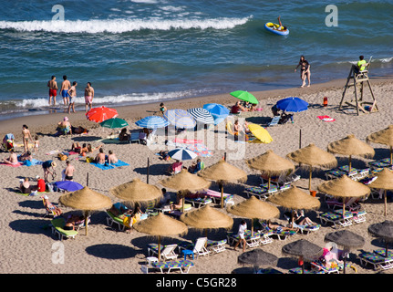 URLAUBER GENIEßEN DAS MEER UND DEN SAND UNTER SONNENSCHIRMEN AM STRAND IN FUENGIROLA COSTA DEL SOL VON EINEM RETTUNGSSCHWIMMER ÜBERWACHT Stockfoto