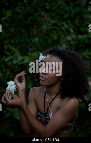 Schöne schwarze Frau mit weißen Blumen im Haar sitzen und draußen einen Badeanzug tragen und lächelnd Stockfoto
