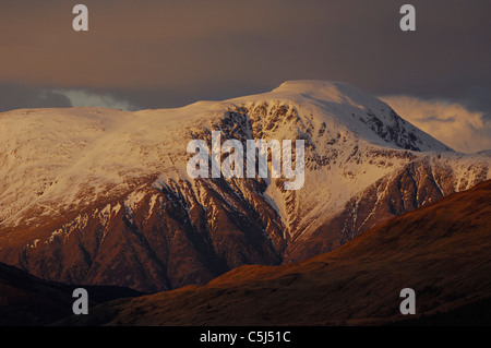 Tele Schuss herzlich beleuchtet, schneebedeckte Gipfel des Ben Nevis, Schottlands höchstem Berg, erhebt sich über dunkle Ausläufer, Stockfoto