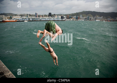 Zwei Männer vor einem Steg am Strand von Freyberg, Oriental Bay ins Meer tauchen. Wellington Stadt im Hintergrund, Neuseeland Stockfoto