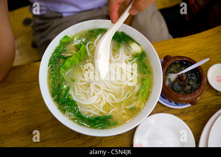 Person / Hand hält einen Löffel über eine Schüssel mit frischer / authentischer / echter chinesischer Nudelsuppe in einem Restaurant in Peking, China. Nudeln scheinen Reis zu sein. Stockfoto