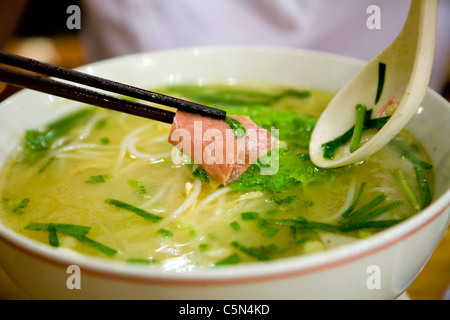 Person / Hand hält einen Löffel über einer Schüssel mit frischer / authentischer / echter chinesischer Nudel- & Fischsuppe in einem Restaurant. Peking, China Nudeln scheinen Reis zu sein. Stockfoto