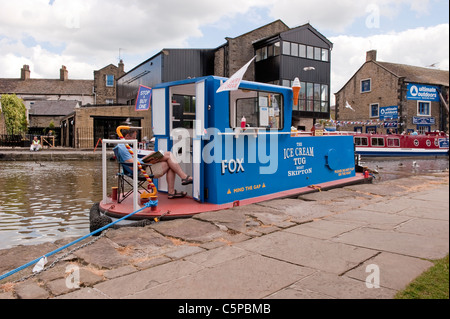 Eisboot 'Fox' (umgebauter Schlepper), das Eis verkauft, das von einem Schlepptau angemacht wurde und der Mann auf dem Deck sitzt, liest - Leeds Liverpool Canal, Skipton, England, Großbritannien. Stockfoto
