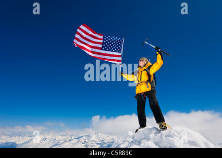 Weibliche Bergsteiger fliegen die Sterne und Streifen auf dem Gipfel des schneebedeckten Berge. Stockfoto