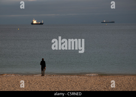 Mann im Mantel stehen am Strand mit Blick auf das Meer auf zwei Schiffe Stockfoto