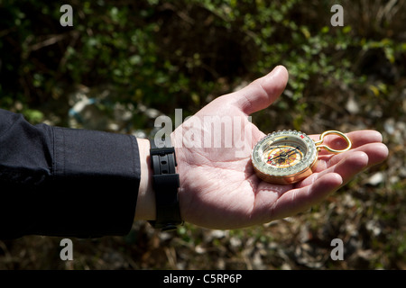 Hand des Mannes hält einen direktionalen Kompass vor einem dunklen bewaldeten Hintergrund. Stockfoto