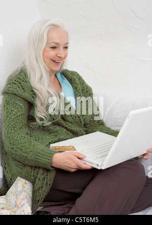 Deutschland, Düsseldorf, Frau sitzt mit Kreditkarte, Karte und Laptop auf Sofa, Lächeln Stockfoto