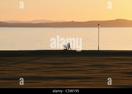 Silhouette von Frauen, die den Kinderwagen bei Sonnenuntergang am Meer in Largs, Ayrshire, Schottland, Großbritannien, schieben Stockfoto