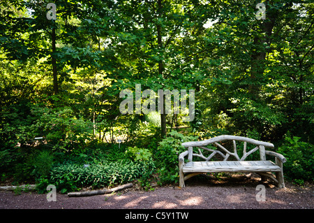Parkbank im US National Arboretum Washington DC | WASHINGTON DC, Vereinigte Staaten — Eine rustikale Bank im Aststil steht neben einem Kiesweg in einer bewaldeten Ecke des US National Arboretums, beschattet von dichtem Sommerlaub. Das Arboretum, ein Forschungs- und Bildungsreservat, das vom US-Landwirtschaftsministerium betrieben wird, verbindet botanische Sammlungen mit ruhigen, naturistischen Gartenbereichen wie diesem. Stockfoto