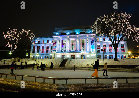 QUEBEC CITY, Kanada – Eisläufer gleiten über eine Eisbahn vor dem eleganten Palais Montcalm. Die beleuchtete Fassade des historischen Gebäudes bietet eine atemberaubende Kulisse für Wintererholung im Herzen des Kulturviertels von Quebec City. Stockfoto