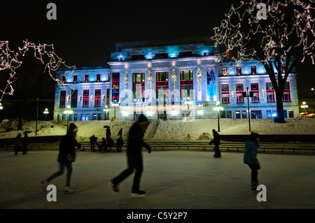 QUEBEC CITY, Kanada – Eisläufer gleiten über eine Eisbahn vor dem eleganten Palais Montcalm. Die beleuchtete Fassade des historischen Gebäudes bietet eine atemberaubende Kulisse für Wintererholung im Herzen des Kulturviertels von Quebec City. Stockfoto