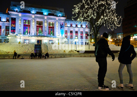 QUEBEC CITY, Kanada – Eisläufer gleiten über eine Eisbahn vor dem eleganten Palais Montcalm. Die beleuchtete Fassade des historischen Gebäudes bietet eine atemberaubende Kulisse für Wintererholung im Herzen des Kulturviertels von Quebec City. Stockfoto