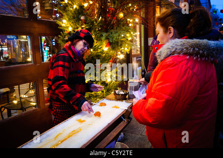 QUEBEC CITY, Kanada – Eine Frau macht einen Ahornsirup-Taffy-Lutscher, indem sie heiße Taffy im Eis auf der Rue du Petit-Champlain in der Altstadt von Quebec dreht, die wunderschön für Weihnachten in der Nacht eingerichtet ist. Die festliche Atmosphäre der Straße macht den Genuss dieses traditionellen Québec Winters noch angenehmer. Stockfoto