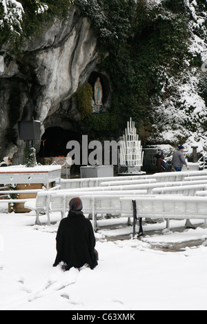 Lourdes im Winter: eine betende Pilger an der Grotte, die Wallfahrtskirche unserer lieben Frau von Lourdes. Stockfoto