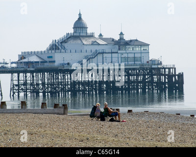 Eastbourne Strand zeigt ein paar relaxen in Liegestühlen mit dem Ende des Piers im Hintergrund am leuchtenden Sommermorgen Stockfoto