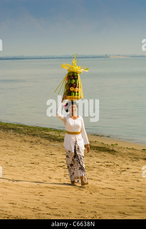 Asiatische Frau in traditioneller balinesischer Kleidung, die an einem Strand mit Obstkorb oder Canang-Sari-Opfergabe auf ihrem Kopf läuft, mit dem Indischen Ozean im Hintergrund. Stockfoto