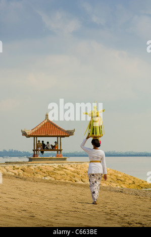 Balinesin in traditioneller Tracht, die am Strand zu einer Pergode geht und einen Korb mit Obst oder Canang Sari auf dem Kopf trägt. Stockfoto