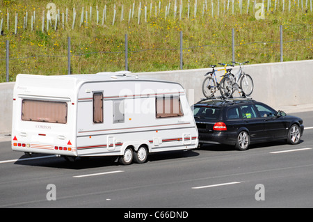 Fahrradfahrer mit Seitenansicht nach hinten auf einem Dachträger auf schwarzem Auto Abschleppwagen Weißer Zweiachser Caravan fährt entlang der Autobahn M25 in Großbritannien Stockfoto