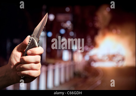 Messerkriminalität / Aufstände / Gewalt Konzept. Die Hand hält ein Zombie-Messer mit einer gezackten Kante, während ein Auto im Hintergrund brennt. London Großbritannien Stockfoto