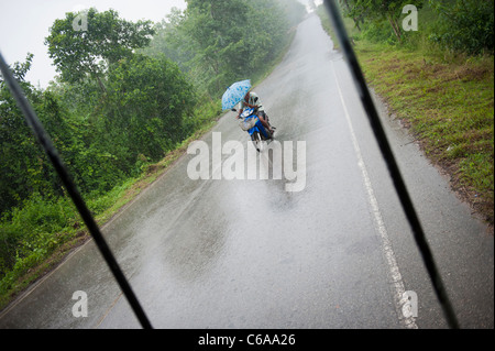 Ein Mann fährt ein Motorrad auf einer Dschungel-Straße bei starkem Regen mit Regenschirm Thailand Asien Stockfoto