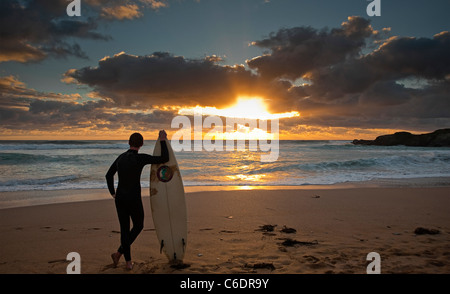 Ein junger Surfer in einen Neoprenanzug steht mit seinem Brett gegen einen spektakulären stürmischer Sonnenuntergang an Konstantin Bay Newquay England Stockfoto