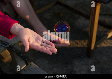 Seifenblase schwebt in Richtung Frauenhand bei Sonnenuntergang, während sie versucht, die Regenbogen-Kugel zu fangen Stockfoto