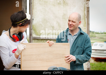 Ein Mann und eine Frau diskutieren die Schleifarbeiten einer Holztür, tragen von persönlicher Schutzausrüstung (PSA) für Sicherheit. Stockfoto