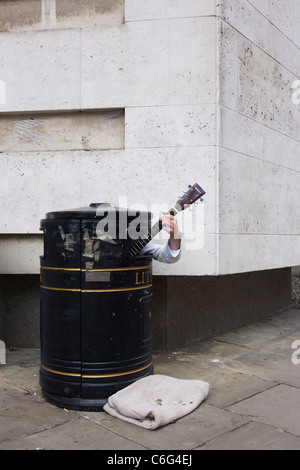 Eine Straße Straßenmusiker versteckt in einem Abfallbehälter, zusätzliches Geld zu verdienen, wie Passanten seine Musik auf einem Cambridge-Pflaster hören. Stockfoto