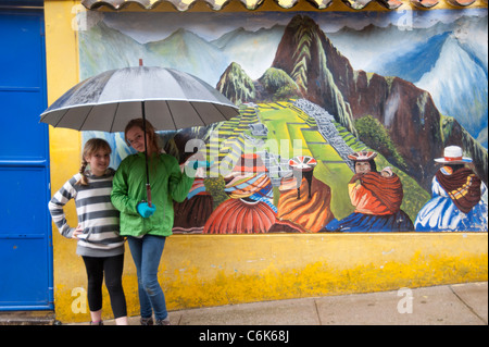 Mädchen, die Zuflucht unter einem Regenschirm vor ein Wandbild von Machu Picchu, Cusco Region, Peru Stockfoto