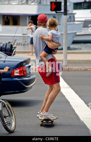 Ein Vater trägt seine Kleinkind Tochter beim Überqueren einer Straße gegen die Ampel auf einem Skateboard in San Diego, Kalifornien. Stockfoto