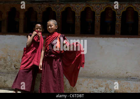 Zwei sehr junge Lehrling Mönche kichern vor einer Bank von Gebetsmühlen im Kloster Wangduephodrang Dzong in Bhutan. Stockfoto