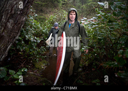 Mann trägt schwere Regenbekleidung hält Surfbrett unter Arm Wandern im Wald Stockfoto