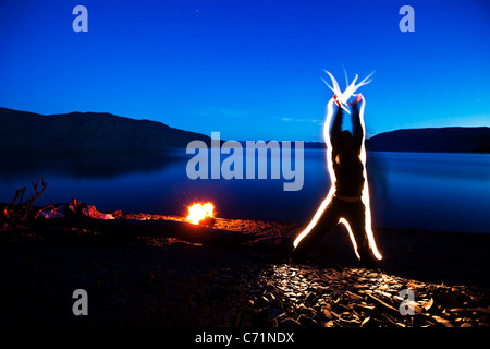 Eine Frau schickt Energie durch ihren Körper und ihre Hände bei Sonnenuntergang neben einem Lagerfeuer und einem See in Idaho. Dieses Licht Malerei imag Stockfoto