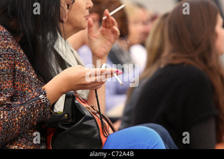 Zwei Damen sitzen in einer Menschenmenge, jede holding eine Zigarette. Stockfoto