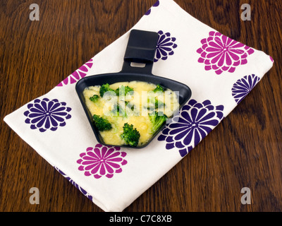 Raclettepfännchen mit Käse und Broccoli auf bunte Serviette Stockfoto