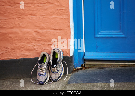 Ein paar alte Trainer lehnten sich im Mai an einer lachsrosa Wand, die mit einer blauen Tür in Padstow, Cornwall, Großbritannien, kontrastierte - sie verließen das Haus Stockfoto