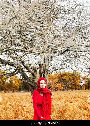 Eine Frau trägt ein leuchtend rotes Kleid und Schal stehen unter einem blattlosen Baum in der Natur Stockfoto