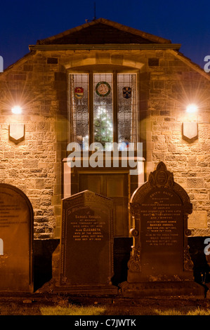 Außenansicht der Abendkirche (Beleuchtung, Buntglasfenster, geschmückte Weihnachtsbaumlichter) und Grabsteine auf dem Kirchhof - Baildon, Yorkshire, England. Stockfoto