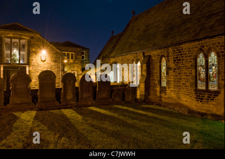 Abendkirche und Grabsteine auf dem Kirchhof (Lichter an, Beleuchtung von Buntglasfenstern und geschmückter Weihnachtsbaum) - Baildon, Yorkshire, England. Stockfoto