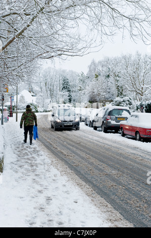 Winter Village Street (schneebedeckte Fahrbedingungen, Autos auf der Straße, Fußgänger zu Fuß, schneebedeckte Bürgersteig) - Burley in Wharfedale, England, GB, Großbritannien. Stockfoto