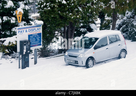 Verschneiten Winter Parkplatz (silbernes Auto im Schnee durch Pay-und Display-Ticket-Maschine geparkt, Liste der Gebühren auf dem Schild) - Baildon, West Yorkshire, England, Großbritannien. Stockfoto