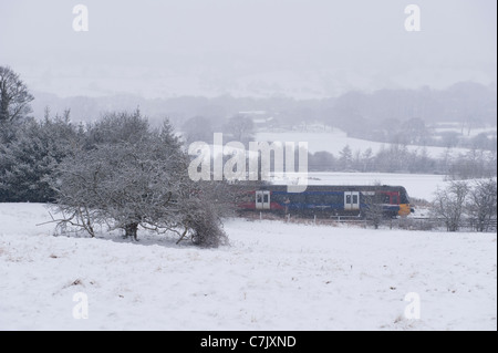 Schnee fällt auf schneebedeckten Wintertag und lokaler Zug der Personenbahn, der an weißen Landfeldern vorbeifährt - Wharfedale Line, West Yorkshire, England, Großbritannien. Stockfoto