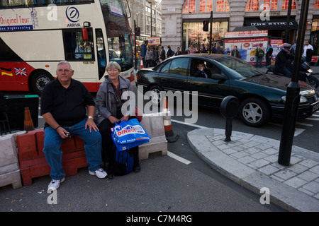 Umgeben von dichten Verkehr, sitzen zwei mittleren Alters Shopper auf eine Straße Barriere in der Mitte des Piccadilly Circus. Stockfoto