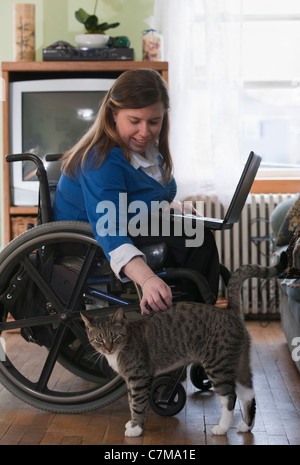 Geschäftsfrau mit Spina Bifida in einem Rollstuhl mit ihrem Laptop zu Hause und ihre Katze streicheln Stockfoto