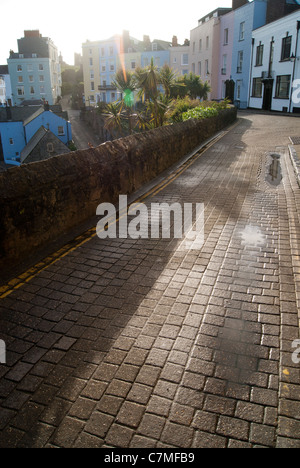 Helle am frühen Morgen gepflasterte Straße mit Pastell farbigen Häusern und Palmen mit Sonneneruption in Tenby, Wales. Stockfoto