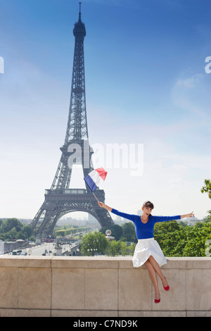 Frau hält eine französische Fahne, sitzen auf einer Steinmauer mit dem Eiffelturm im Hintergrund, Paris, Ile de France, Frankreich Stockfoto