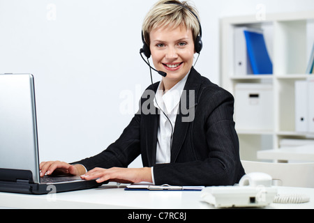 Junge hübsche Rezeptionistin mit Kopfhörer am Computer Blick in die Kamera und lächelnd Stockfoto