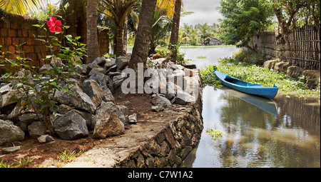 Landschaft von Kerala Backwaters Indien Arterie festgemacht blau Kanu lokalen Stein Granit Findlinge Hibiskus Strauch mit einsamen Blüte Stockfoto