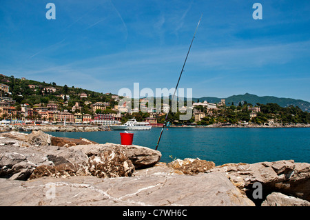 Einen roten Eimer auf den Felsen und eine Angelrute erscheinen von hinten gegenüber die intelligenten Gebäude von Santa Margherita Ligure, Italien Stockfoto