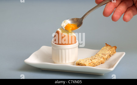 A mans Hand löffelt einen Teil des weich gekochtes Ei mit gerösteten Getreidespeicher "Soldier" auf einer kleinen Platte Stockfoto